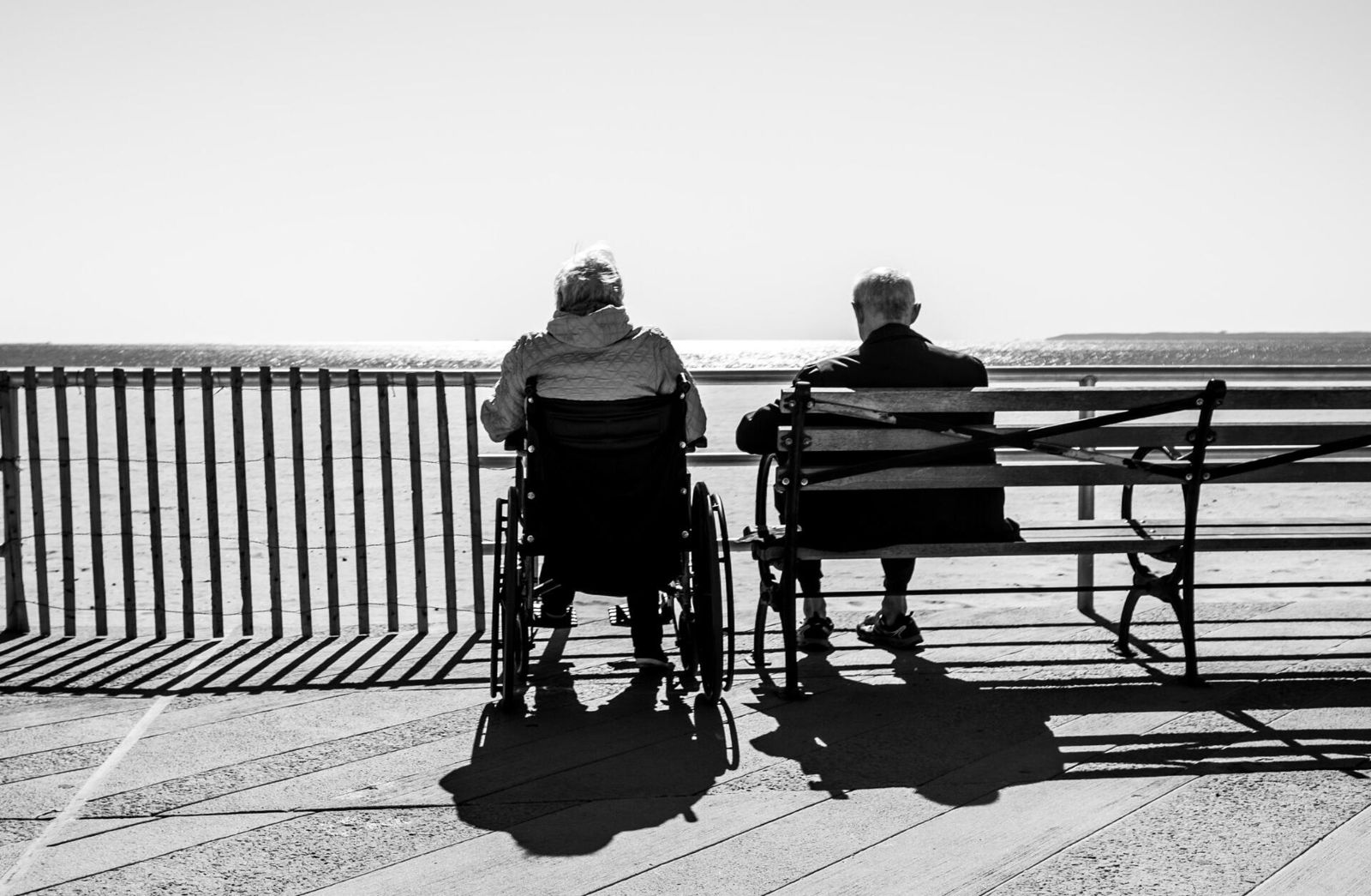 An older couple looking out over the water from a boardwalk, one in a wheelchair, the other on a bench beside them.